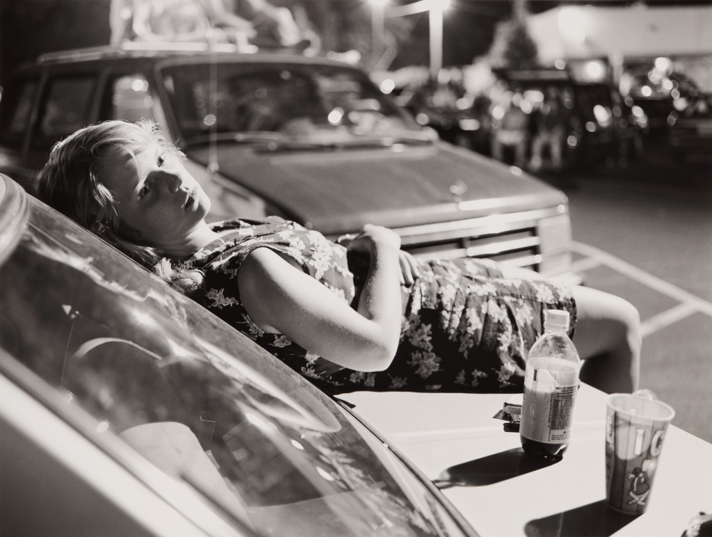 Mark Steinmetz An adolescent girl with short blonde hair reclines on the hood of a car, a bottle of soda and a paper cup filled with ice sit next to her.