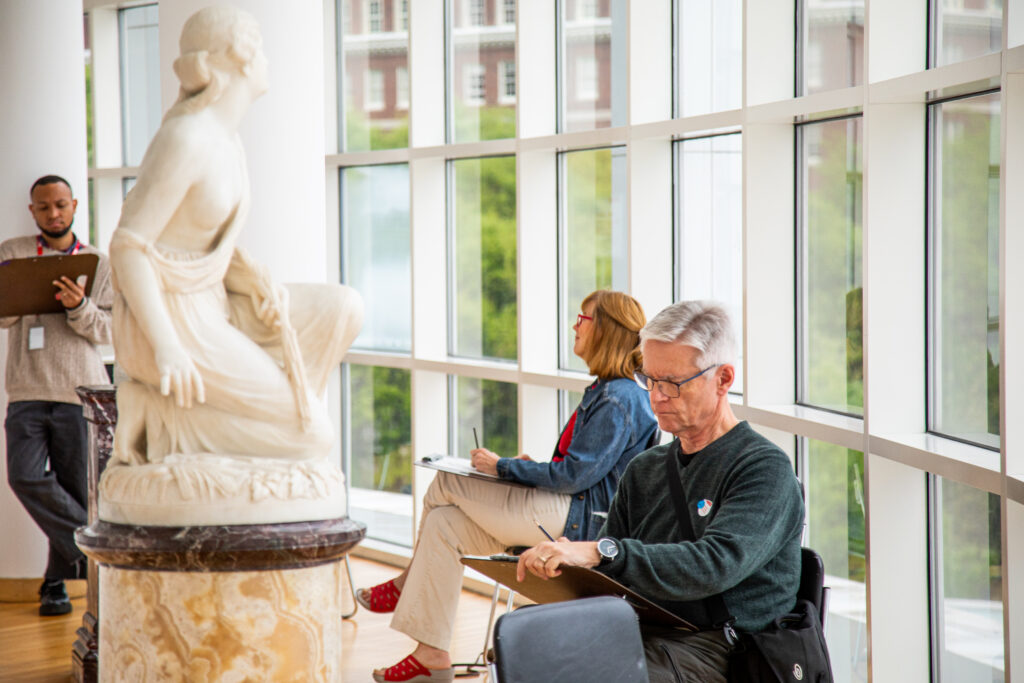 One person stands while two other people sit while sketching a marble sculpture in front of them