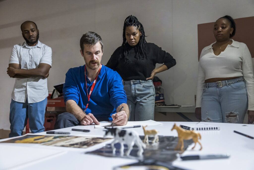Art instructor is sitting down and drawing while students stand behind watching closely