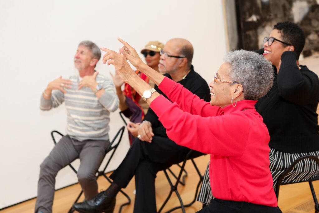 older woman with brown skin and grey hair points both hands at a painting as she explains to a group how it makes her feel.