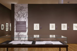 The Plot Thickens installation shot - a case with prints sits in front of an installation of King Lear prints.