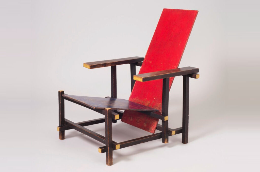 Two wooden planes, painted red, form the seat and back of this simple chair with wooden arms that run parallel to the floor and perpendicular to the legs.